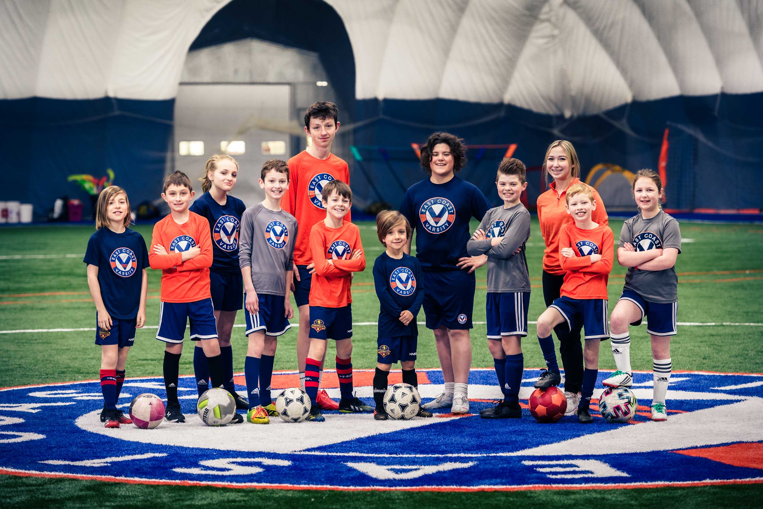 A group of students of different ages with soccer balls on the turf.